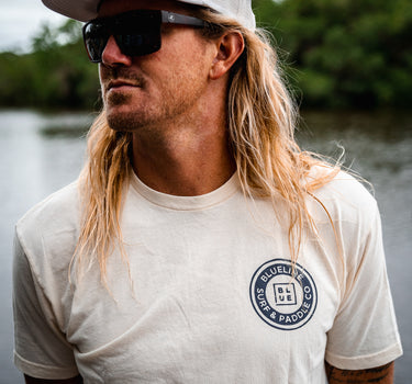 Man wearing a washed natural Blueline Surf & Paddle Co. T-shirt with a navy OG circle logo, standing near the water in Jupiter, Florida.