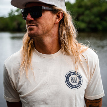 Man wearing a washed natural Blueline Surf & Paddle Co. T-shirt with a navy OG circle logo, standing near the water in Jupiter, Florida.