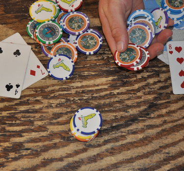 Hand holding Blueline poker chip style golf ball markers on a wooden table with playing cards and chips in the background.