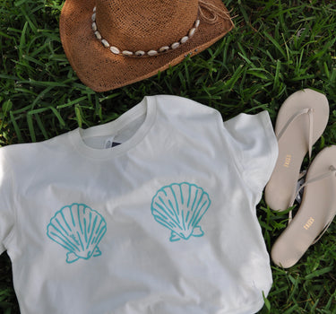 Women's Blueline Shell Crop Tee in natural featuring a coastal shell graphic, styled with a straw hat and sandals on the grass.