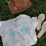 Women's Blueline Shell Crop Tee in natural featuring a coastal shell graphic, styled with a straw hat and sandals on the grass.