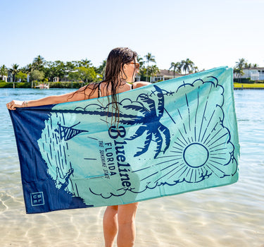 Person holding a Blueline Sunshine State beach towel with palm tree and Florida graphics while standing in shallow ocean water.