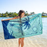 Person holding a Blueline Sunshine State beach towel with palm tree and Florida graphics while standing in shallow ocean water.
