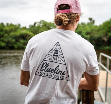 Man wearing a washed white Blueline Triangle Lighthouse T-shirt with navy graphic on the back, standing on a dock near the water in Jupiter, Florida.