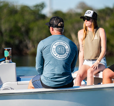 Man sitting on a boat wearing a heather slate blue Blueline long sleeve shirt with a white Vintage Circle logo on the back.