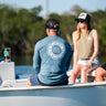 Man sitting on a boat wearing a heather slate blue Blueline long sleeve shirt with a white Vintage Circle logo on the back.