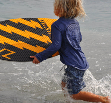 Child wearing the Blueline Youth OG Logo Hooded UV Sun Shirt in navy while carrying a skimboard into the ocean for a day at the beach in Jupiter, Florida.