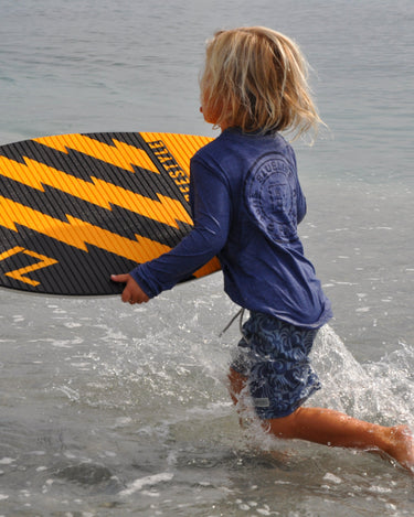 Child wearing the Blueline Youth OG Logo Hooded UV Sun Shirt in navy while carrying a skimboard into the ocean for a day at the beach in Jupiter, Florida.