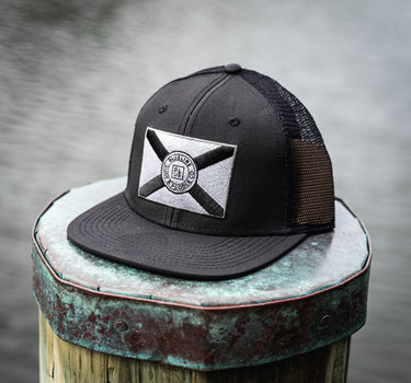 Black Blueline trucker hat featuring a blacked-out Florida state flag patch, photographed on a dock near the water in Florida.