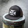 Black Blueline trucker hat featuring a blacked-out Florida state flag patch, photographed on a dock near the water in Florida.