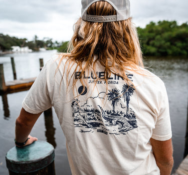 Man wearing a washed natural Blueline Gator Lasso T-shirt with light navy artwork while standing on a dock near the water in Jupiter, Florida.