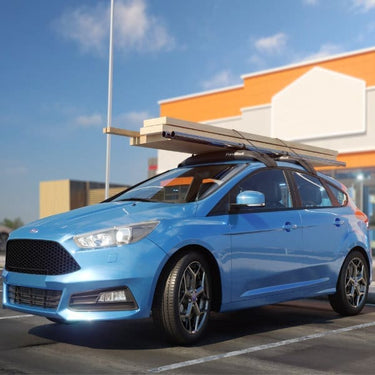 Side profile of a Ford Focus sedan with lumber stacked and secured on the HandiRack temporary roof rack, ready for construction.