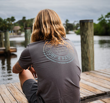 Man sitting on the docks at Guanabanas in Jupiter, FL wearing a blueline asphalt shirt back view.