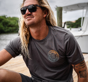 Man wearing a dark asphalt gray Blueline short sleeve T-shirt with an orange and blue faded OG Blueline logo while sitting on a dock near the water.