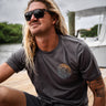 Man wearing a dark asphalt gray Blueline short sleeve T-shirt with an orange and blue faded OG Blueline logo while sitting on a dock near the water.