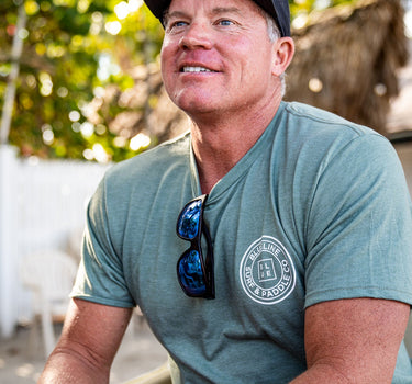 Man wearing a canyon green Blueline OG logo T-shirt with white chest logo, seated outdoors near the beach in Jupiter, Florida.
