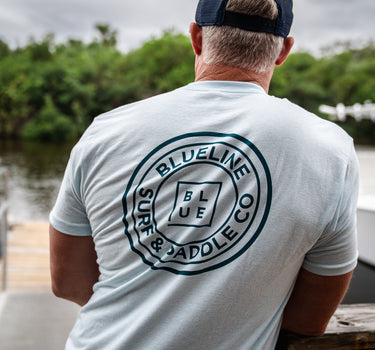 A person wearing a navy and light blue t-shirt with the text 'BLUE LINE SURF CO' on the back, standing near a body of water.