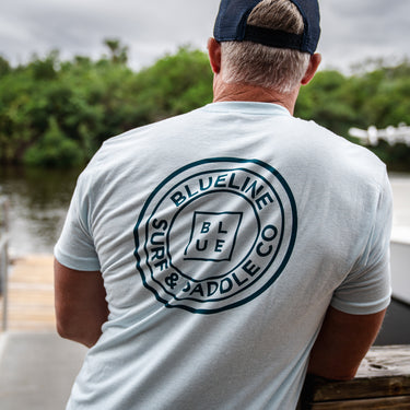 A person wearing a navy and light blue t-shirt with the text 'BLUE LINE SURF CO' on the back, standing near a body of water.
