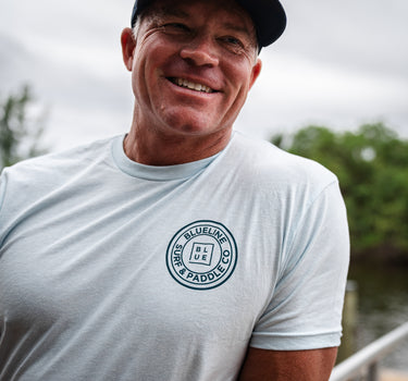 Man wearing an ice blue Blueline short sleeve T-shirt with a navy OG Blueline Surf & Paddle logo while standing near the water in Jupiter, Florida.