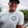 Man wearing an ice blue Blueline short sleeve T-shirt with a navy OG Blueline Surf & Paddle logo while standing near the water in Jupiter, Florida.