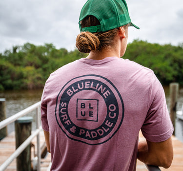 A person wearing a navy t-shirt with the text 'BLUE LINE SURF & PADDLE CO.' on the back, standing near a body of water.