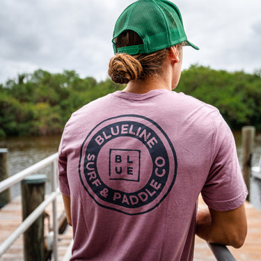 A person wearing a navy t-shirt with the text 'BLUE LINE SURF & PADDLE CO.' on the back, standing near a body of water.