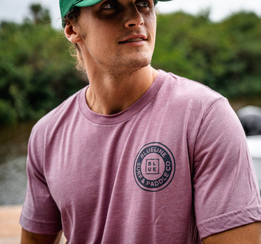 Man wearing a light orchid Blueline short sleeve T-shirt with a navy OG Blueline logo on the chest and back while standing near the water.