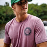 Man wearing a light orchid Blueline short sleeve T-shirt with a navy OG Blueline logo on the chest and back while standing near the water.