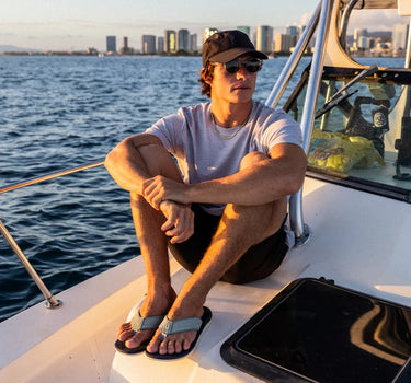 Man wearing OluKai ʻAhi Kai sandals while relaxing aboard a boat at sunset