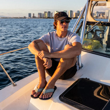 Man wearing OluKai ʻAhi Kai sandals while relaxing aboard a boat at sunset