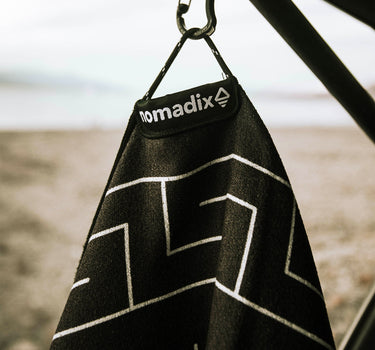Black towel with white zigzag pattern hanging from a hook on a blurred beach background
