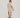 A woman viewed from the back wearing a short-sleeve button-up shirt dress in a light beige and white floral pattern, paired with brown strappy sandals, against a plain light gray studio background.