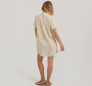 A woman viewed from the back wearing a short-sleeve button-up shirt dress in a light beige and white floral pattern, paired with brown strappy sandals, against a plain light gray studio background.