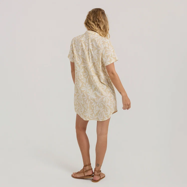 A woman viewed from the back wearing a short-sleeve button-up shirt dress in a light beige and white floral pattern, paired with brown strappy sandals, against a plain light gray studio background.