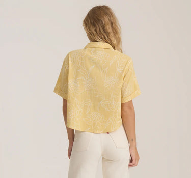 A woman viewed from behind wearing a pale yellow short-sleeve button-up shirt with a white floral line pattern, paired with off-white pants, against a plain light background.