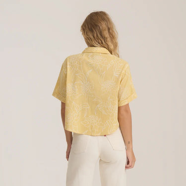 A woman viewed from behind wearing a pale yellow short-sleeve button-up shirt with a white floral line pattern, paired with off-white pants, against a plain light background.