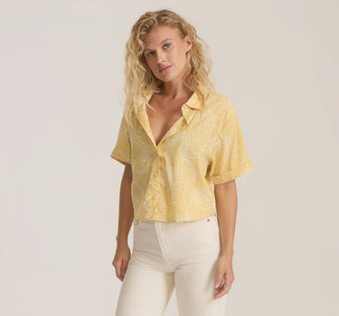 Woman wearing a yellow patterned blouse and white pants on a plain background