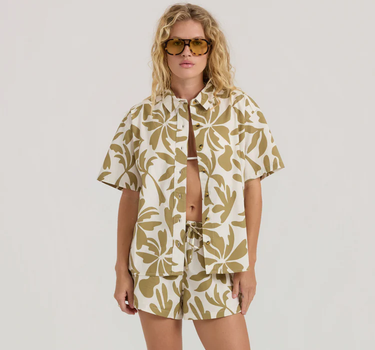 Woman wearing a beige and brown patterned shirt and shorts set with sunglasses on a plain background