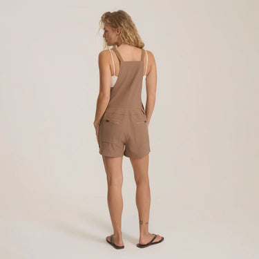 A woman stands with her back to the camera wearing a taupe-brown sleeveless romper with thin shoulder straps and cargo-style back pockets, paired with dark flip-flop sandals, against a plain light beige studio background.