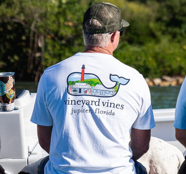 Man sitting on a boat wearing a white Vineyard Vines and Blueline T-shirt with a Jupiter Lighthouse storefront graphic on the back.