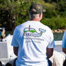 Man sitting on a boat wearing a white Vineyard Vines and Blueline T-shirt with a Jupiter Lighthouse storefront graphic on the back.